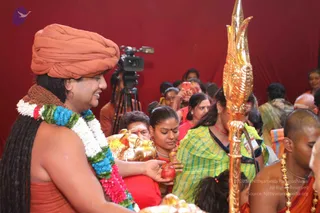 A photograph of a crowded darshan scene against a red backdrop. In the left foreground a figure in an orange saffron turban and an orange robe wears a multicolour flower garland of white, green, blue and pink blossoms and a brown rudraksha-style necklace, smiling as he reaches out to hand a red apple to a woman. Beside him a woman in a green and yellow striped sari holds a red apple, and a gold ceremonial vessel is visible below. On the right a golden trident-like staff stands upright, and a bald man in an orange garment wears gold beads. A video camera on a tripod and a few devotees holding up phones and watching from behind fill the background. Probable Swamiji (orange turban, long swept-back hair, flower garland and rudraksha necklace) is handing out fruit as a blessing.
