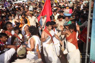 A dense crowd fills a street during a procession, with many people pressing forward and waving. In the foreground, several men wear white dhotis with gold borders and orange-red shawls, some bare-chested with rudraksha and bead necklaces, and they gesture with raised hands as they move. A man in an orange top and a shaved-head figure in white stand among them. Uniformed police officers in khaki stand along the route, one holding a radio. A woman in a green sari carries a child, and a red flag rises above the crowd at right.