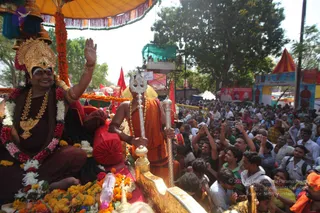 A festival procession on a decorated golden palanquin-style chariot beneath a yellow canopy with orange marigold garlands. A figure seated on the left wears a gold crown, a dark maroon robe, white and pink flower garlands, and gold necklaces, and raises one hand in greeting. Beside him a standing attendant in saffron-orange robes and rudraksha beads holds a tall silver trident. Dense crowds press along the road, many raising mobile phones, and red flags and tented stalls line the street under green trees.