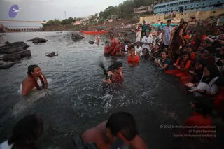 Devotees bathe in the Narmada river at Omkareshwar during a snaan at dusk. In the foreground a man with wet hair and a red garment is bent forward in the water, splashing, while a bare-chested man on the left smiles with a beaded mala around his neck. Behind them a crowd stands waist-deep and along the rocky bank, many in orange and red clothing, some in white. A man in a white shirt and a woman in a white sari stand on the steps. Stone ghats, riverside buildings, a bridge and a red boat fill the background.