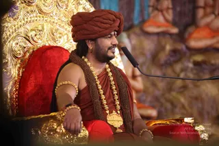 A man seated on an ornate gold-and-red throne with a red velvet cushion, speaking into a microphone on a black stand at right. He wears a rust-red turban, a rust-red robe draped over his bare shoulder, a brown rudraksha-style mala, a heavy gold ceremonial necklace with a temple-pendant, gold bangles on his arms, and a gold earring. Behind him, blurred painted or sculpted figures in orange robes sit in meditation poses against a rocky backdrop. a person: long hair, ornate gold necklace and malas, and red-orange clothing with a turban.