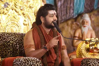A man seated on a leopard-print cushioned chair in front of an ornate gold throne-style back panel, speaking into a black microphone while pointing with his right index finger. He wears a rust-orange wrap over the shoulders, a long brown beaded rudraksha-style mala, orange beaded armlets, a red bracelet and a gold earring, with white and red forehead markings. Beside him stands a painted or sculpted figure of an elderly white-bearded sage with hands folded in prayer, and a gold ornamental object is visible at the lower right. The scene resembles a discourse in progress. Appearance cues are consistent with the page's a person label, with a rust-orange wrap, rudraksha mala and ornate gold setting, so the candidate is probable.