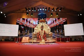 A photo of a stage set for a discourse. A man in orange-red robes, a red and orange turban, and a long white and gold garland sits cross-legged on an ornate gold throne with a black crown-like canopy and lion-carved arms. Gold trident-style staffs (tridents) stand on either side of the throne, and a red carpet covers the floor. Two whiteboards flank the stage: the left one shows a diagram of the 4 states of consciousness with terms like Jagrat, Swapna, Sushupti and Turiya; the right one is labelled 'The 25 States of Consciousness' with a table of Sanskrit-transliterated terms. A large backdrop shows a red-and-white colonnaded hall. Stage lights hang from a truss above. The seated figure is an appearance-based a person in a throne-seated pose with rudraksha-style garland and orange robes, though the image is a single seated presenter on stage.