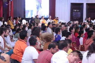 A large seated audience fills a hall during a satsang, facing a screen at the top left that shows a bearded man in saffron and white with a turban and ornate necklace. Many attendees wear white, orange and saffron clothing; several men wear rudraksha malas. Women in yellow, pink and blue sit on white chairs near the front, and a man in white with a mala sits at the left edge. The room has red curtains and tall wooden doors.