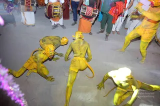 A night procession scene on a bare ground with several men painted entirely yellow, wearing yellow loincloths, yellow face masks with fierce features and long tails, crouching and leaping in a dynamic dance pose. Behind them, drummers in white dhotis and orange cloth stand playing large drums with red and white woven covers, while onlookers in green and white stand watching. A purple flower garland is visible at the left edge.