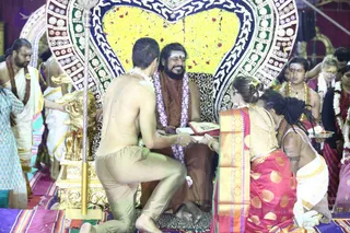 A seated bearded man in a brown-orange robe and a white-and-purple flower garland sits smiling on a leopard-print cushioned throne in front of a large yellow flower arrangement with red petals. A shirtless man in beige trousers kneels at left, holding out a plate, while a woman in a red and gold sari kneels at right offering items on a tray. A brass lamp stand with an ornate golden base stands in the foreground. Other people in white dhotis and garlands, one in turquoise, surround the scene on a striped floral mat. The bearded seated man shows a distinctive ceremonial figure with a garland and throne, with cues limited to his brown robe and flower garland, so identity is uncertain.