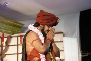 A photograph of a man seated on a stage with his palms pressed together in anjali mudra and his eyes closed. He wears a rust-red turban with a small silver ornament at the front, a pink and white lotus-petal garland, a red-orange cloth over his shoulders and across his torso, and rust-red beaded bracelets and armlets. Long dark hair falls past his shoulders and a brown rudraksha-style mala hangs on his chest. Behind him are a green-gold ornate panel, a white backdrop and red and white hanging garlands, with a microphone clipped at the bottom edge. Possibly a person by appearance, with several cues: long swept hair, a crown-like turban and orange ornate clothing.