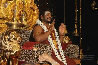 A man seated on an ornate gold throne with leopard-print cushions and a golden lion armrest, palms joined in greeting and smiling. He wears a red-brown robe, a white-and-blue flower garland, gold armbands and bangles, and a white forehead marking. A brass kalasha-style lamp and bells sit on the left, with a small oil lamp held by an unseen hand at lower left. Gold ornaments and string lights decorate the dark background. a person seated in a darshan setting; the throne, leopard-print cushions and golden lion armrest are visible, and the figure wears rudraksha-style gold adornments with a red-brown robe and garland.