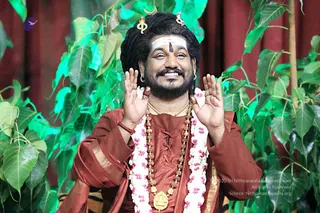 A smiling man with long dark matted hair topped by a gold crescent ornament and a gold trident-like hair piece stands before green peepal leaves and a maroon curtain. He wears a rust-red saffron-toned robe with gold trim, a white and pink jasmine garland, multiple rudraksha malas, and a large gold ceremonial pendant. A white tripundra with a central dark mark decorates his forehead, and he raises both palms in a greeting gesture.