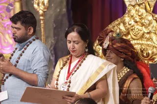A stage event photograph with a golden ornate throne and a golden deity-style figure in the background. In the foreground, a woman in a white saree with a gold border and a red lanyard, wearing a rudraksha mala, looks down while writing on a wooden board labelled GURUKUL. To her left, a bearded man in a light blue shirt also wears a rudraksha mala and looks down at a paper. Behind her, a man in a maroon turban with a peacock feather ornament, gold necklaces and a maroon garment stands near the throne, and a microphone is held at the right edge. Three physical people are visible in the scene.