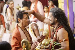 A photograph of a ritual moment in an ornate hall. On the right, a long-haired man with flowing dark hair, a beard, and a bare chest wears a maroon-brown cloth, layered rudraksha malas, a gold ceremonial necklace, a rudraksha bracelet and an armlet; he presses his right hand to the forehead of a shorter man with short dark hair seated on the left. That seated man wears an orange-saffron shawl printed with text over bare shoulders, a rudraksha mala, and a gold chain, and keeps his eyes closed with his hands raised in reverence. Behind them, several people in white dhotis and orange shawls stand and sit, a boy in an orange shawl is visible, and a golden deity murti sits on the lower centre near a small oil lamp and a brass vessel. White tripundra marks are visible on a seated man's arm.