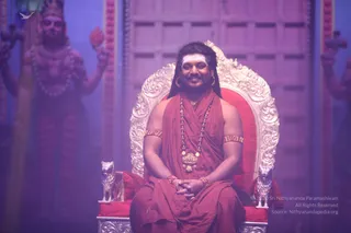 A man seated on an ornate silver-and-gold throne with red cushions, flanked by silver lion figures at its base. He wears a red-orange robe draped over his shoulders and bare chest, a long red-orange rudraksha-style bead mala, a gold ceremonial pendant necklace, gold bangles on his arms and wrists, and gold hoop earrings. His long dark hair is swept back, and he sits with hands folded in his lap, smiling. Behind him, pink-lit deity murti statues with raised arms stand against a wooden panelled wall. The scene is lit in purple and pink tones. Swamiji (probable) is seated on the throne, wearing saffron-red robes, rudraksha mala and ceremonial gold necklace, with long swept-back hair.