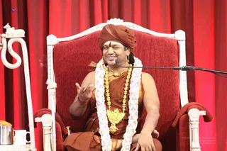 A man seated on an ornate white and red throne chair in front of a bright red draped backdrop, speaking with his right hand raised in a gesture while a microphone stand is positioned beside him. He wears a rust-orange turban, a rust-orange wrap, a long white flower garland, and a heavy gold necklace with rudraksha-style beads and a gold pendant. A small red mark is on his forehead, and a cup and a silver tumbler sit on a small table at the left, with a carved white naga-style armrest visible at the left edge. Swamiji (probable): the rust-orange turban and wrap, long white garland, gold ceremonial necklace and seated discourse setting match the cues for the speaker at this event.