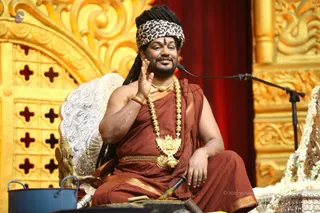 A photograph of a man seated on an ornate golden throne with carved gold panels and a white silver-patterned backrest. He wears a leopard-print headband, a rust-brown robe with gold border, a heavy gold ceremonial necklace with a large gold pendant, and a red bracelet, and has a red tilak on his forehead. He raises one hand in a blessing gesture while a microphone stands beside him. a person appears here based on the long dreadlocked hair, ornate gold necklaces, and saffron-brown ceremonial clothing. A blue bucket and a small ritual object sit at the lower left. Red curtains and white flower garlands frame the stage.