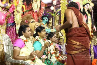 Night photograph of a Navaratri puja at a temple with large painted deity murtis wearing gold ornaments and garlands. In the foreground, three women sit on a silver-coloured chair holding garlands of white and orange flowers, with a trident and a pink lotus visible. The woman in the centre wears a teal blouse and a rudraksha mala, while the woman on the left wears a pink blouse and cream sari. Behind them, a bare-backed man in a rust-red dhoti and shawl stands facing the deities, holding a brass bell in his raised hand. A small child in a crown and ceremonial dress stands at the right edge. Garlands and a lion-headed figure are visible among the decorations.