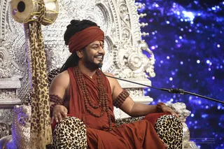 A smiling man sits on an ornate silver throne with carved floral and figural details, a leopard-print cushion beneath him. He wears an orange-red turban, an orange-red shawl over his shoulders, multiple rudraksha malas around his neck and arms, and a small tilak on his forehead. A microphone on a stand is in front of him, and a leopard-print fabric hangs from the throne on the left. The backdrop is a deep blue starry sky. Swamiji (probable) is seated in the Navaratri darbar, with long swept-back hair, an orange-red turban and rudraksha malas. No other people are visible.