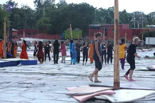 Outdoor scene on a stone-paved courtyard where many people practise Shivasthamba (mallakhamba) pole yoga around upright wooden poles. Participants in orange, maroon, black, blue and yellow clothing stand and move near the poles; several wear black athletic clothes and sandals. In the foreground a woman in a black sleeveless top and dark capris walks past a pole, and an older woman with short blonde hair in black clothes and pink shoes stands at right. Folded mats lie on the ground, and a green generator truck, red-walled buildings and trees appear in the background.