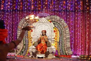 A photograph of a seated man on an ornate silver throne with a dark-patterned embellished backrest, in a decorated stage set with string lights and draped curtains in purple, red and gold. He wears a red-orange turban, orange-saffron robes and a gold-coloured ceremonial necklace, and raises his hands in a blessing gesture. In the left foreground a person's arm holds a lit oil lamp, its flames glowing. Marigold garlands hang on the throne, and brass lamps line the stage floor. Visible appearance cues include a red-orange turban, saffron robes and gold ornaments, so the seated figure is a probable Swamiji (probable) presiding in person, seated on the throne during an arati.