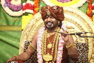 A photograph of a smiling man seated on an ornate gold throne with a leopard-print cushion, speaking into a microphone on a stand. He wears a rust-brown turban decorated with a jewelled ornament and a small gold emblem, a rust-brown robe, a heavy gold necklace and multiple strands of gold beads, gold armlets and bangles, and long garlands of pink lotus-like flowers. Behind him are bright marigold and rose garlands and green draped cloth. A copyright-style credit line appears in the lower right corner of the photo.
