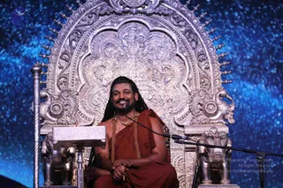 A man with long dark hair and a beard sits smiling on an ornate silver throne with a large repoussé backrest and elephant-head armrests, in front of a starry blue backdrop. He wears a rust-orange wrap over his shoulders, a gold-toned beaded mala and a long gold necklace. He rests his hands on his lap and sits beside a white lectern with a microphone on a stand. Swamiji (probable): the long swept-back hair, orange ceremonial wrap and gold necklaces on a silver throne match the page's claim, though the face is not used for identification.