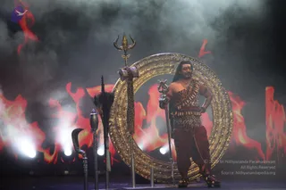 A photograph of a stage performance set against a dark smoky backdrop with red and orange flame-like projections. A single standing man in a stylised Shiva-like costume rests one hand on his hip while holding a tall silver trident topped with a trident finial. He wears a leopard-print bodice with a dark red and maroon wrap, a beaded belt, a rudraksha-style necklace and bead strands across his chest, and dark footwear. Beside him stands a large golden circular ring-shaped prop with ornate carved edges, along with a dark staff with a curved top and a tall spear-like pole topped with a dark feathered tuft. Bright stage lights glow at the base of the scene.