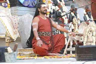 A seated man in a rust-red sleeveless garment and matching drawstring trousers sits cross-legged on a colourful woven mat before a silver-carved throne. He has long dark dreadlocks, a white horizontal forehead mark, white ash markings on his arms, and a leopard-print waist sash with a rudraksha mala draped over his shoulder. He rests one hand on his knee and holds a white staff, eyes closed. Behind him stand dark deity murti statues draped with marigold garlands, a brass lamp stand and an ornate brass-and-silver peetha, with a microphone on a stand at the right. a person based on long dreadlocks, ash markings, rudraksha mala and ceremonial sash.