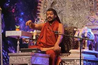 A seated man in saffron-orange robes gestures with his right hand while speaking into a microphone on a stand. He wears a red-orange tilak on his forehead, a red thread on his wrist, a long braided hairstyle and a small necklace. He sits in an ornate silver throne with a leopard-print cushion and carved silver elephant armrests. A silver side table holds a green-and-orange water bottle and a Yamaha DTX device. The backdrop is a starry galaxy image in blue and purple. Swamiji (probable): the cues are long swept-back hair, saffron robes, ornate silver throne and a seated discourse setting, so this is a teaching or satsang-style talk.