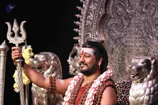 A bearded man sits on an ornate silver throne with a repousse back, flanked by silver lion sculptures. He wears a saffron-red cloth over his bare shoulder, layered rudraksha malas, a gold chain and pink-and-white flower garlands, with a white horizontal forehead mark and red bindu. He holds a silver trident in his raised right hand, with yellow flowers hanging beside it, and smiles toward the left against a black backdrop. Probable Swamiji: long swept-back hair, rudraksha malas, saffron clothing and a seated ceremonial throne with a silver trident.