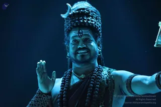 A performer in a blue-lit stage setting wears a tall matted headdress of rudraksha beads topped with a silver crescent, with a white tripundra and a bindi-like mark on the forehead. He wears layered rudraksha malas, a gold chain and bead armbands, and a dark draped cloth with a patterned shawl. He smiles and raises one palm while extending the other arm, holding a lantern at the right edge. The styling evokes Shiva iconography, with no visible event text or other people.
