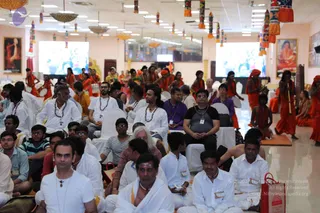 Photograph of a seated audience in a large hall with cream walls, hanging colourful tassel decorations and round lamps overhead. Many seated attendees wear white clothes and rudraksha malas, and some wear name badges. In the foreground a bearded man in a white shirt and a woman in purple sit among them. On the right, saffron-robed performers with orange turbans play drums and long wind instruments near a stage, and two large screens show footage behind them. Portraits hang on the walls, and a red-and-white bag sits on the floor.