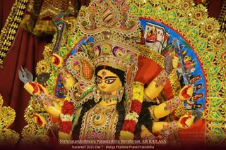 A richly decorated Durga deity murti with a golden crown and ornate embroidered jewelled costume, shown with multiple arms holding a trident, a sword, a disc and other weapons. Red and white flower garlands hang at its shoulders and a red bindi marks the forehead. Behind it is a golden arched halo with a painted blue and red backdrop. The murti is installed at Nithyanandeshwara Paramashiva Devalayam, Adi KAILASA during Navaratri Day 7, a Durga Pratima Prana Pratishtha. No people are visible.