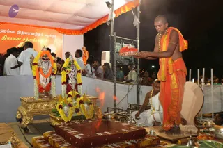 Night outdoor ritual under a white tent with orange trim. In the foreground a shaved-headed priest in orange dhoti and orange shawl with beaded necklaces stands holding a brass plate with red flowers, facing two decorated deity murtis with silver crowns, flower garlands and orange drapes on a gold platform. Beside him a younger attendant in white dhoti and white shawl kneels near a red-brown homa or yagna fire pit with a flame and brass vessels. Behind the stage, a crowd of devotees watches, some in white and some in colourful saris. A banner with Tamil script stands at left, and a flower-covered offering tray sits at lower right.