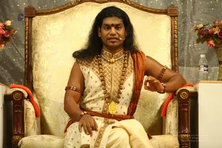 A man with long dark hair sits on an ornate gold-brocaded armchair with carved wooden arms and orange cushions. He wears a cream embroidered sleeveless kurta with a rust-orange shawl draped across one shoulder, a rudraksha mala and a large gold pendant, and cream dhoti-style trousers. A red bindi marks his forehead, and he wears a headset microphone while speaking, one hand resting on his knee. Pink and orange flowers sit in a vase to the right, a water bottle stands beside him, and a sparkling grey backdrop hangs behind. Swamiji (probable) appears as the seated speaker, identified by long swept-back hair, ornate gold-embroidered clothing, rudraksha mala and gold pendant. The image is a photo of a discourse setting.