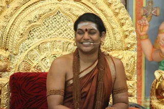 A smiling man sits on a red tiger-print cushioned seat in front of an ornate gold throne with intricate carved backrest. He wears a rust-brown and gold-bordered shawl over a bare chest, layered brown rudraksha malas, and gold armlets. He has a white tripundra and red bindi on his forehead and a small headset microphone near his mouth. Beside him a painted figure of a deity with raised hands holds a trident.
