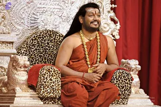 A man with long dark hair and a short beard sits on an ornate silver throne with lion-head armrests and a leopard-print cushioned seat, set against a red curtain. He wears a rust-orange sannyasi wrap over his shoulder and draped cloth, a heavy gold bead necklace with a gold pendant, and a red tilak on his forehead with a white marking above it. He smiles with his hands resting in his lap. Swamiji (probable) is identified by long swept-back hair, a saffron-orange wrap and an ornate gold necklace, seated on a throne.