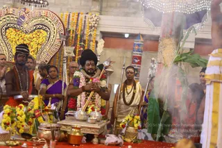 A photograph of a Hindu ritual inside a temple hall decorated with marigold and orange garlands. At the centre, a man with a tall dark turban-like hairstyle, a red tilak, a flower and rudraksha-style garland, and a saffron-and-white ceremonial cloth holds a ritual staff with both hands before a small silver altar stand with brass pots and offerings. Smoke rises at the right near a tall decorated pillar. Around him stand attendants: a young man in saffron with bead necklaces holding a metal bowl, a woman in a purple sari, a bald man in white with a rudraksha mala holding a silver staff, and other devotees in saffron and white. A large ornate golden arch with a decorated canopy frames the back.