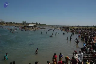 A wide outdoor photograph of a crowded river bathing ghat at a Kumbhamela-style festival, with hundreds of devotees wading and bathing in shallow, muddy-green water under a clear blue sky. Many people in saris and dhotis stand in the water or along the sandy bank, some in red, pink, yellow and green garments. Thatched and tin-roofed shelters and a row of bamboo stalls line the far bank beside leafy trees. Dense crowds fill the right foreground. No identifiable a person figure is visible in the scene.
