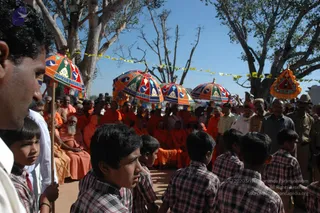 Outdoor photo at a temple-town event under bare trees and a clear blue sky. Several large umbrellas with multicoloured panels and tassels, printed with a dove-like emblem and the words 'Dhyanapeetam', are held up over a seated group of saffron-robed sannyasis and devotees on sandy ground. A bearded elder in orange sits at left, and a man in a white shirt stands at the far left holding an umbrella pole. In the foreground, several boys in checked shirts face away or look toward the group. Policemen in khaki uniforms and a man in a hat stand at right among the onlookers, and strings of small yellow and white flags hang across the trees. No one is clearly identifiable as a person in this image.