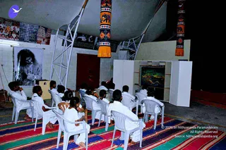 A group of about fifteen people in white clothes sit on white plastic chairs on a colourful striped woven rug inside a tent-like hall with a white canopy and tall pillars wrapped in orange and black elephant-patterned fabric. On the left, a large black-and-white portrait of a long-haired man in a dark robe with hands clasped is mounted on a stand, and banners with photographs hang along the upper wall. On the right, a white screen-like frame displays a colourful painting. Several attendees face the front, and the room is dimly lit.