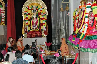 A temple puja in progress before a central deity murti draped in green, yellow and red garlands, with a gold crown and gold-framed arch behind it. A seated priest in saffron-orange robes with a shaved head sits at the lower right, facing the deity with hands folded, beside an offering tray holding apples, a bowl and a small arrangement of red flowers. On the left, a man in orange clothing kneels with palms pressed together, and another devotee in orange sits beside him. Devotees sit in rows on the floor in the foreground, with red rope barriers between them and the shrine. Two brass bells hang at the upper right, and a large second deity murti in a pink, green and gold skirt with garlands stands at the right edge.