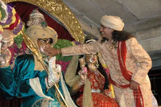 A photograph of a wedding-style ritual at a temple. A man in a cream embroidered sherwani and matching cream turban, with long dark hair and a red sash draped over his shoulder, reaches out to touch the forehead of a golden metal murti of a deity. The murti wears a teal velvet garment, a patterned turban with a peacock feather, a golden trident-like staff, and beaded ornaments. Beside it, a second golden murti in a red embroidered dress and gold jewellery holds a pink flower garland and a lotus-shaped object. Behind them are garlands of marigolds and other flowers in purple, pink and green, and a red curtain with gold decorative framing. The man appears to be a probable Swamiji, identified by his long hair, cream turban and ornate embroidered clothing, shown in person at the ritual.