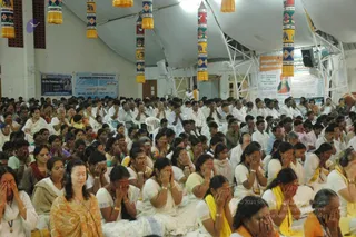 A large seated congregation fills a tented hall at a Kailasa event, with hundreds of devotees sitting on white chairs and mats. Most wear white kurtas and dhotis or saris in white, yellow, pink and orange, and many cover their eyes with their palms in meditative prayer. A woman in the front left wears an orange-and-cream sari with headphones, eyes closed. Hanging decorative pillars with yellow tassels and painted elephant motifs line the ceiling. Banners on the walls carry printed text and a photograph, and a man in white stands near the back. No a person candidate is clearly visible in the frame.