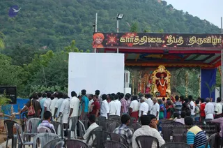 Outdoor photo of a crowd gathered in front of a decorated stage at the foot of a wooded hill. Dozens of people sit on white and grey plastic chairs, facing a shrine-like stage with red and gold draping and a gilded canopy. On the stage sits a deity murti in orange cloth with a garland, flanked by brass lamps and a small kalasha-like arrangement. Several standing devotees in white dhotis and shirts, women in saris, and children fill the foreground and middle. A large banner above the stage shows a smiling figure in an orange turban and Tamil text. A blank white screen stands to the left, and a portrait-style board is visible at the far left edge.