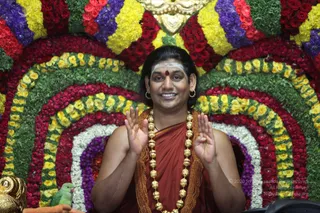 A smiling man sits in front of a dense backdrop of concentric arches made of red roses, yellow and purple flowers and green foliage. He wears a saffron-orange robe draped over bare shoulders, a long necklace of large gold beads, gold hoop earrings, a white vertical tilak and a red dot on his forehead. He raises both palms outward in a blessing gesture. A small green parrot perches on the left edge beside a gold ornament.