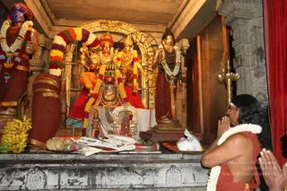 A temple shrine with a deity murti of a seated figure in a gold crown, adorned with gold necklaces and draped in garlands of white jasmine and multicoloured flowers. Two standing figures flank the central deity, one with a crown and one bearing a turban, along with a gilded arch behind. Bananas and offerings sit on a stone platform in front. A person in orange-red robes and a white flower garland stands at the right with palms pressed together in prayer, facing the shrine. Brass lamp stand visible at right.