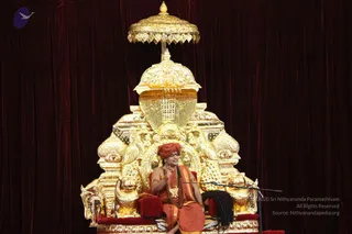 A photograph of a seated man in saffron-orange robes and an orange turban, wearing a gold-toned chain and a rudraksha mala, seated on an ornate gold throne with lion carvings and a golden canopy against a deep maroon velvet backdrop. He raises one hand in a teaching gesture before a microphone stand. Long dark hair falls behind the throne. The scene shows a single speaker on stage with no other people visible.