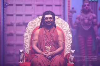 A man with long dark hair swept back and a white forehead mark sits on an ornate silver-and-gold throne with red cushioning, flanked by two small silver lion figures at its base. He wears a red-orange saffron wrap over his shoulders, a rudraksha mala with ornate gold pendant, gold armlets and beaded bracelets, and holds his hands folded in his lap. Behind him, a large blurred deity murti in red with a crown, necklaces and a raised arm stands against a pink-lit wooden panelled backdrop. a person is seated in a ceremonial setting with a throne and deity statue.