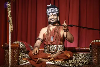 A photograph of a seated man in saffron-orange dress with a leopard-print headwrap topped by a silver crescent, reclining on a carved wooden leopard-print-cushioned seat before a deep red curtain. He wears multiple rudraksha malas, a silver pendant, bangles and armlets, gold-edged saffron cloth around the torso and waist, and raises one index finger while speaking into a microphone on a stand. A tall silver-topped staff with a leopard-print tassel stands to his left. Swamiji (probable): long dark hair, ornate saffron clothing, rudraksha malas and ceremonial gold ornaments are visible on the seated speaker, who is giving a discourse.
