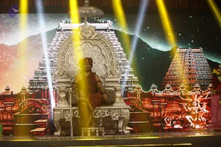 A man in saffron-orange clothing and a red-orange turban sits cross-legged on an ornate silver throne with a leopard-print cushion on a stage. Golden spotlight beams cut through the scene. Behind him is a large backdrop of illuminated gopuram temple towers beneath a starry night sky, with decorative temple sculptures at the lower corners. A silver parasol-style canopy stands above the throne, and a small silver offering vessel sits near the base. Probable Swamiji is seated on the throne, wearing a saffron turban and orange ceremonial clothing, with a bare upper torso and arm ornaments. Nearby on the right, two seated figures appear in meditation on the backdrop artwork, not as physical people. Flower garlands hang at the right edge.