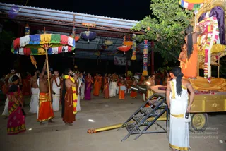 Night procession scene at an outdoor temple-style courtyard under a corrugated roof strung with colourful paper lanterns and bunting. On the right, a golden palanquin-style chariot with a golden canopy and a garland is decorated with flowers; a man in an orange garment stands on it, and a woman in orange stands beside him. A metal ladder leads up to the vehicle. Left, a man in white dhoti holds a multicoloured fringed umbrella over a group. A bare-chested man with a saffron garland, and attendants in white and orange robes, some playing drums, gather in the courtyard. A woman in a magenta sari stands at left. A long golden pole lies on the ground.
